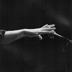 Photographie de Roland Einhaus utilisée sur le site des Abattoirs pour faire la promo de son exposition. Elle est en noir et blanc. L'arrière plan est sombre. Sur la droite de l'image, un micro sur pied se devine, sortant de l'ombre. Un avant bras surgit de la partie gauche de l'image. Presque nu à l'exception d'un bout de chemise sombre, il vient placer sa main au centre de l'image. Le majeur en effleure à peine le micro, les autres doigts tenus, comme en suspension.