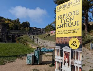 Photo de l'entrée de l'expérience Explore Lyon Antique. Au fond, on y voit beaucoup de verdure. Des arbres et une colline se détache en arrière plan. Un chemin en pierre, qui part d'en bas, grimpe le long de la colline, parré de barrières. Au début du chemin, un panneau pédagogique vert ; à côté d'un grand panneau vertical sur lequel, au dessus de la photo d'un bâtiment antique, un bandeau jaune pâle indique "Lugdunum: musée théâtres romains - EXPLORE LYON ANTIQUE - une expérience immersive au coeur de Lugdunum." Un badge de la même couleur indique également "50 ans".