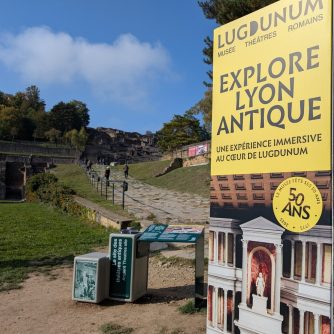 Photo de l'entrée de l'expérience Explore Lyon Antique. Au fond, on y voit beaucoup de verdure. Des arbres et une colline se détache en arrière plan. Un chemin en pierre, qui part d'en bas, grimpe le long de la colline, parré de barrières. Au début du chemin, un panneau pédagogique vert ; à côté d'un grand panneau vertical sur lequel, au dessus de la photo d'un bâtiment antique, un bandeau jaune pâle indique "Lugdunum: musée théâtres romains - EXPLORE LYON ANTIQUE - une expérience immersive au coeur de Lugdunum." Un badge de la même couleur indique également "50 ans".