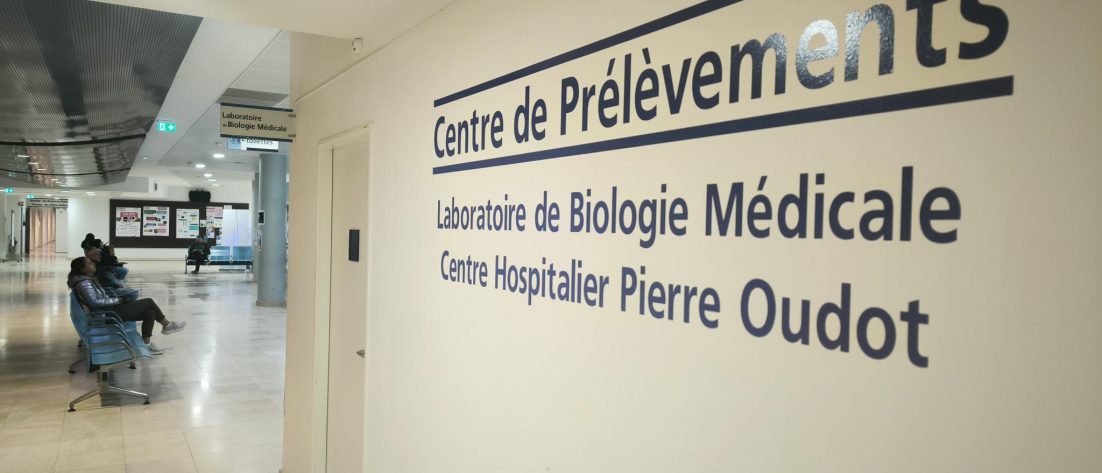 Section du centre hospitalier, dans le hall. Sur la droite de l'image, un mur au bout duquel on aperçoit une porte. Sur le mur est indiqué "Centre de prélèvements" puis "Laboratoire de biologie médicale" et enfin "Centre hospitalier Pierre Oudot".