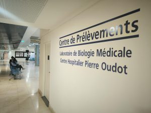 Section du centre hospitalier, dans le hall. Sur la droite de l'image, un mur au bout duquel on aperçoit une porte. Sur le mur est indiqué "Centre de prélèvements" puis "Laboratoire de biologie médicale" et enfin "Centre hospitalier Pierre Oudot".