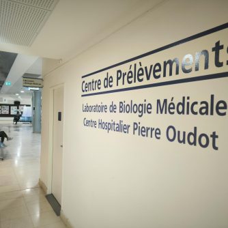 Section du centre hospitalier, dans le hall. Sur la droite de l'image, un mur au bout duquel on aperçoit une porte. Sur le mur est indiqué "Centre de prélèvements" puis "Laboratoire de biologie médicale" et enfin "Centre hospitalier Pierre Oudot".