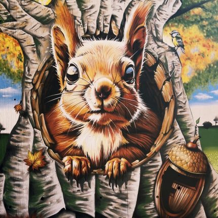 Photo du détail d'une fresque à Bourgoin-Jallieu. On y voit, peints, un immense chêne sur lequel est perché un petit oiseau bleu (peut-être un geai), et duquel sort un énorme écureuil. Aux pieds du chêne, un gros gland ouvert qui ressemble à un tonneau. En bas à gauche de l'image, une signature assez peu lisible.