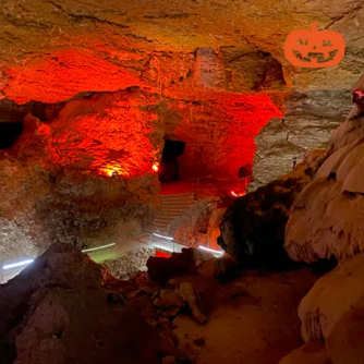 Une photo de l'intérieur des Grottes de la Balme lors d'une précédente édition du Balmoween. On y voit les roches formant des chemins escarpés, de petites galeries étroites, une citrouille peinte sur l'une des façades, le tout éclairé de rouge donnant une ambiance légèrement horrifique.