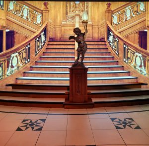 Immense escalier de bois aux rampes sculptées. Il se scinde en deux parties arrivé au premier pallier, et est posé sur un sol carrelé. En son centre, une rampe en bois se clôturant par une statue d'angelot tenant une torche dans ses mains.