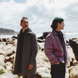 Visuel de tournée pour Sula Bassana. En fond, une falaise, des roches, le tout surplombant l'océan. Le ciel est couvert de quelques nuages blancs. Légèrement à gauche du centre, le saxophoniste Christophe Panzani. Il porte un sweet noir et un épais manteau gris qui lui arrive en haut des cuisses. Légèrement à droite du centre, le producteur 20syl. Il a le regard tourné vers l'horizon et les mains dans les poches de son pantalon. Il porte un bonnet vert foncé, un pull bleu marine et une veste en jean mauve.