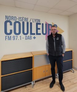 JP Barret pose dans le studio de Radio Couleurs. C'est un homme basique, aux cheveux courts gris, barbichette, lunettes. Il porte une parka noire par dessus un pull gris, un pantalon lui-même noir et des chaussures en daim asorties.
Il pose devant un meuble, posé devant le logo de Couleurs.